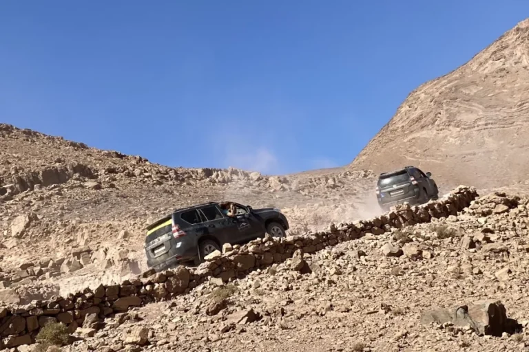 Dos todoterrenos suben por un camino rocoso de montaña bajo un cielo azul despejado. Detrás de los vehículos se forma una estela de polvo.