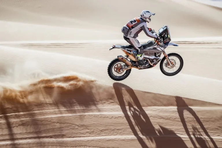 A motorcyclist rides across sandy dunes, kicking up dust, under a clear sky.