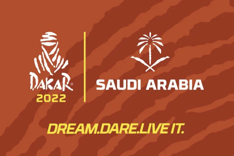 Dakar 2022 logo with a stylized figure, Saudi Arabia emblem, and text: "DREAM. DARE. LIVE IT." against a brown striped background.