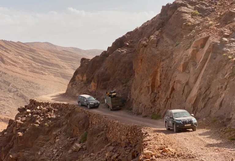 Tres coches circulan por una carretera de montaña estrecha y rocosa con acantilados escarpados bajo un cielo parcialmente nublado.